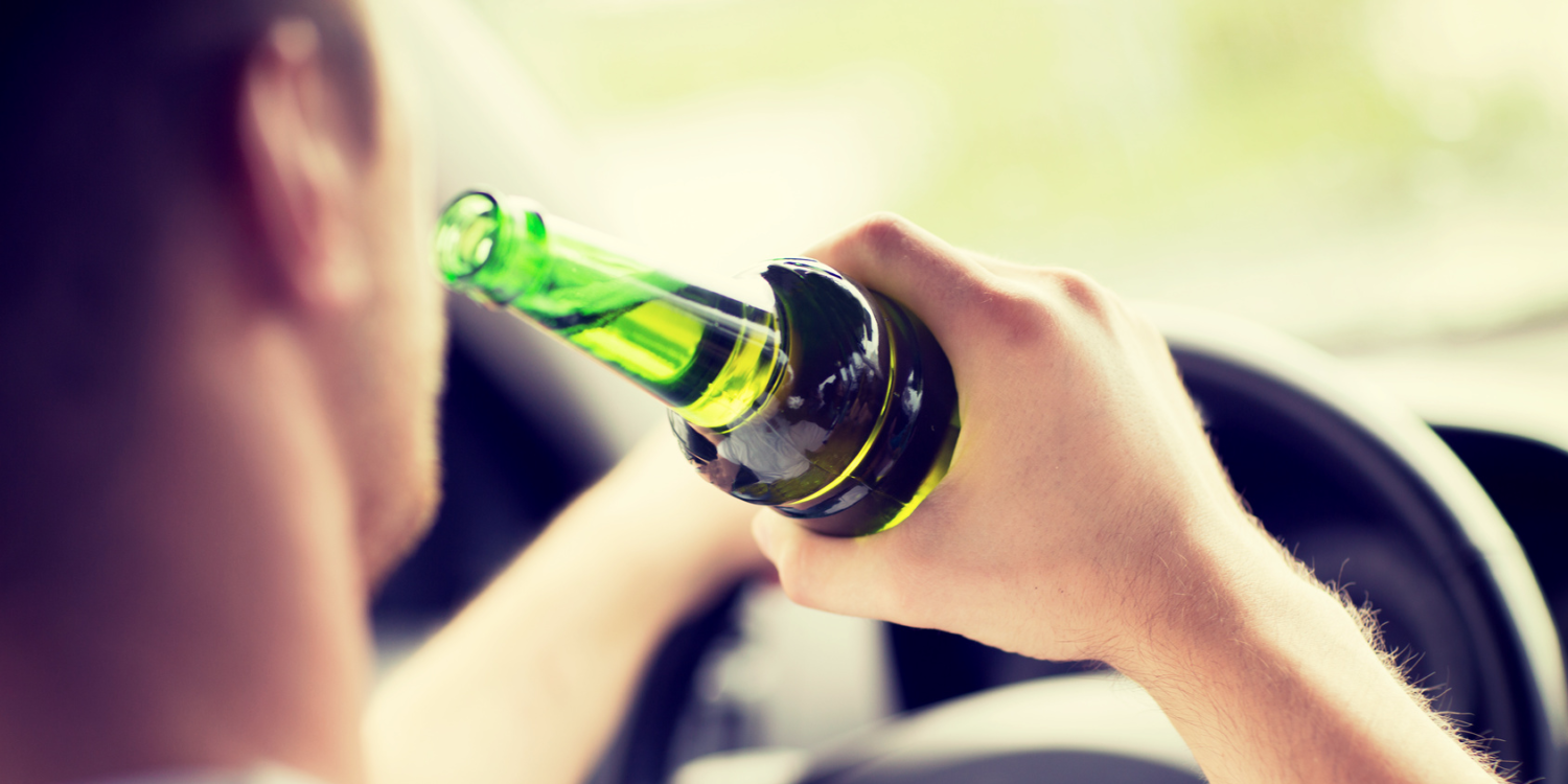 homem ingerindo bebida alcoolica enquanto esta dirigindo, segurando a garrafa com a mão direita e dirigindo com a mão esquerda, dirigindo durante o dia, garrafa na cor verde e volante na cor preta. 