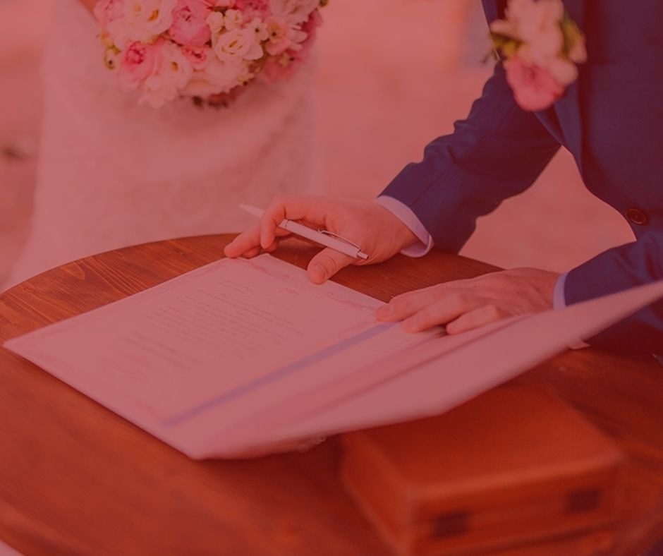imagem com fundo vermelho. Artigo: A importância do pacto antinupcial. Na foto, duas pessoas assinando acordo de casamento, ela vestida de noiva com um vestido branco, ele vestido de noivo com um terno azul. Acordo esta sendo assinado sob uma mesa de madeira na cor marrom.