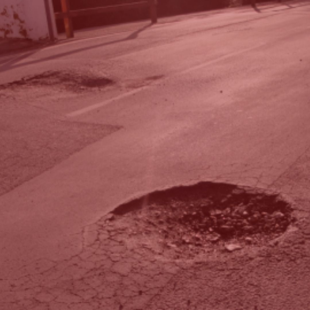 Notícia: Prefeitura indenizará mulher que se acidentou em buraco na via. Na foto, uma rua com dois buracos no meio das vias. Cores na foto: vermelho e cinza.