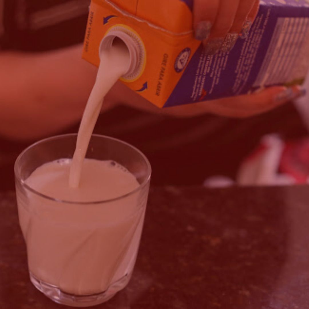 notícia: Procon Notifica Fabricantes de Laticínios Por Aumento no Preço do Leite. #Pracegover: Na foto, uma pessoa colocando o leite em um copo. Cores na imagem: laranja. azul, preto e verde. Fundo vermelho.