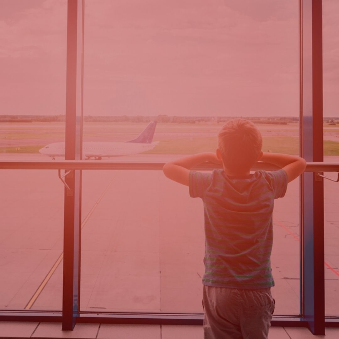 notícia: Crianças serão indenizadas por companhia aérea após pernoite em aeroporto. #pratodosverem: na foto, uma criança em um aeroporto olhando para um avião que está prestes a decolar. Cores na foto: vermelho, verde, azul, branco, cinza a preto. 