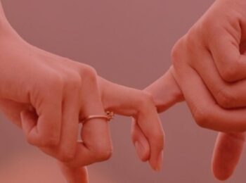 artigo: Possibilidade de reconhecimento de união estável após a morte. #pratodosverem: na foto, um casal segurando o dedo um do outro, com a aliança da noiva a mostra. Cores na foto: vermelo, cor de pele e dourado.