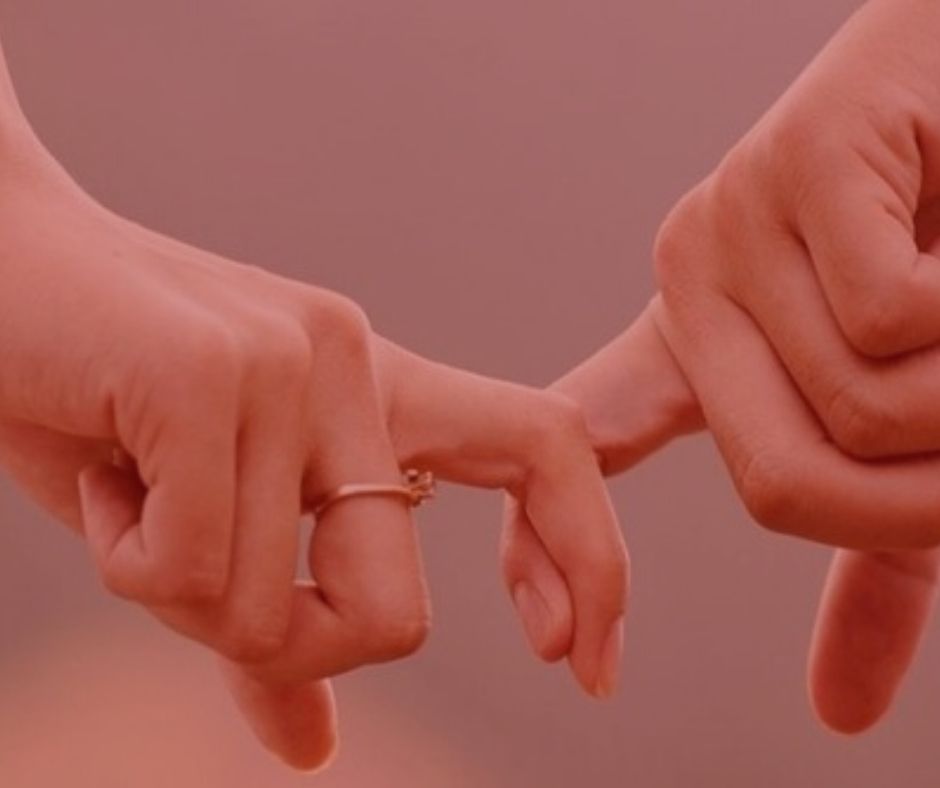 artigo: Possibilidade de reconhecimento de união estável após a morte. #pratodosverem: na foto, um casal segurando o dedo um do outro, com a aliança da noiva a mostra. Cores na foto: vermelo, cor de pele e dourado. 